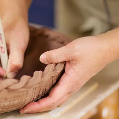 Set in a pottery studio, the image is a close up view of hands holding a detailed clay bowl. Hatching it with a tool.