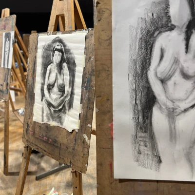 Images of two easels, with charcoal drawn pieces depicting a nude woman with black hair, no facial features and her hands held in front of her.