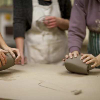 An image of three people in a pottery studio, their faces out of view with a focus on two peoples hands kneading clay at a table.