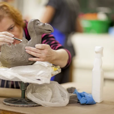 Set in a pottery studio, a woman delicately carves details onto a large clay bird sculpture with it in the foreground.