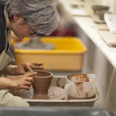 A person is sat at a pottery throwing wheel in a studio. They have their hands on the clay, sculpting it into a bowl-like shape.
