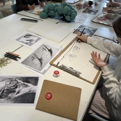A young person is drawing hands using their own as a reference. The table they are seated at is covered in pictures of hands.