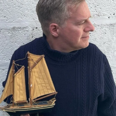 Person in a dark blue knitted jumper holds a small wooden sailing ship model with tan sails, against a pale brick wall.