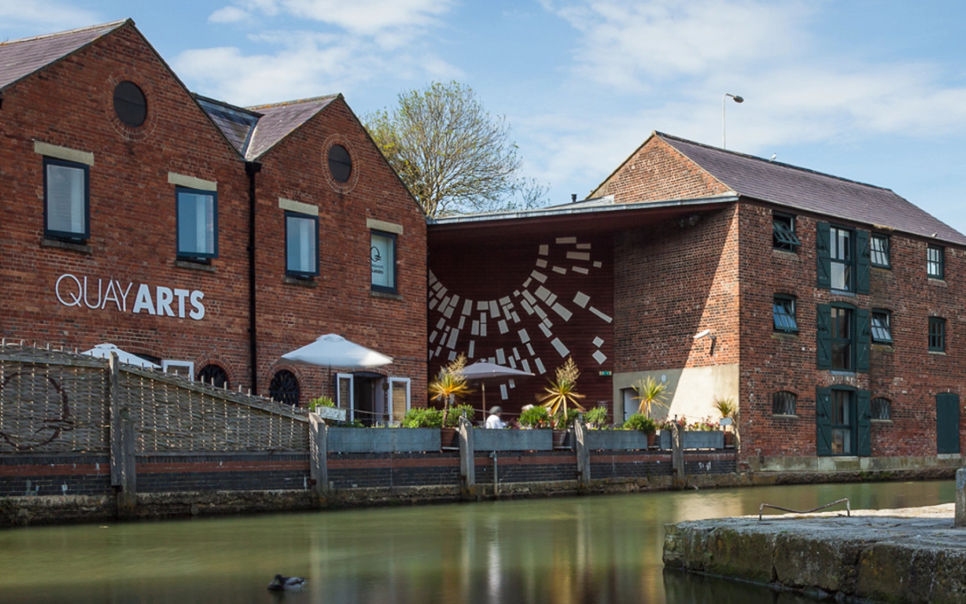 Red brick buildings by a canal, Quay Arts sign, outdoor seating with umbrellas, and a white geometric mural on the central wall; a duck on the water.